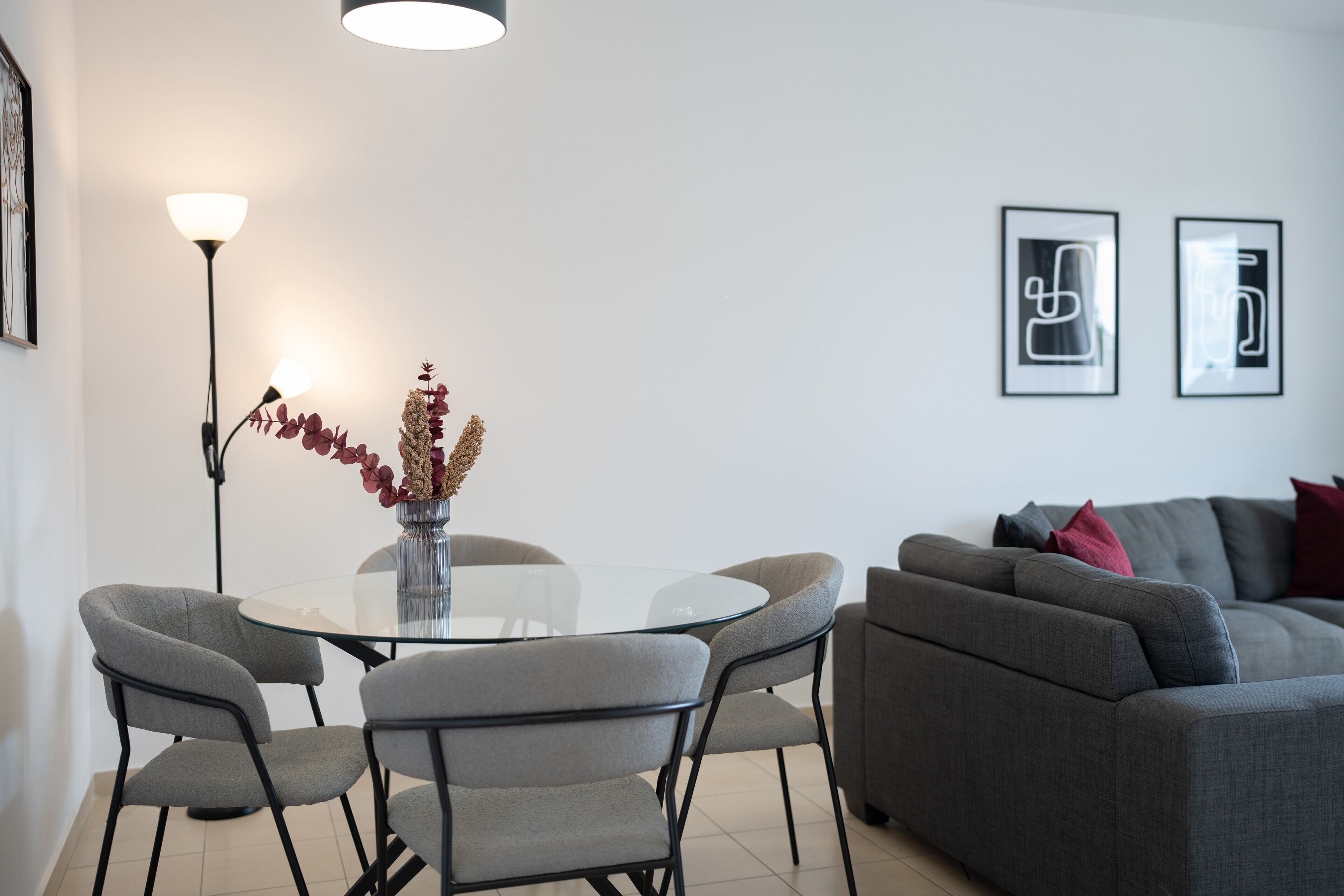 A modern living room with a glass dining table, grey sofas, and a floor lamp.