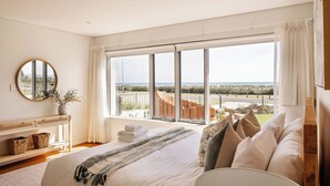 Een slaapkamer met een groot raam, een bed met kussens, een nachtkastje en een spiegel.