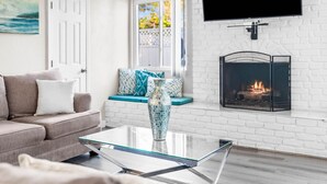 A living room with a glass coffee table, a beige sofa with patterned cushions, a fireplace, and a TV mounted on the wall. - Tranquil Escape Pool Hot Tub Game Room (San Diego)