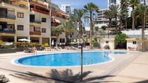 A swimming pool surrounded by lounge chairs and palm trees, with multi-story buildings in the background.