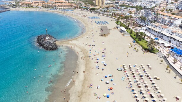 A beach with a rocky outcrop, numerous umbrellas, and a variety of buildings in the background.