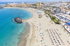 A beach with a rocky outcrop, numerous umbrellas, and a variety of buildings in the background. - Los Cristianos. Fantastic seafront apt (Arona)