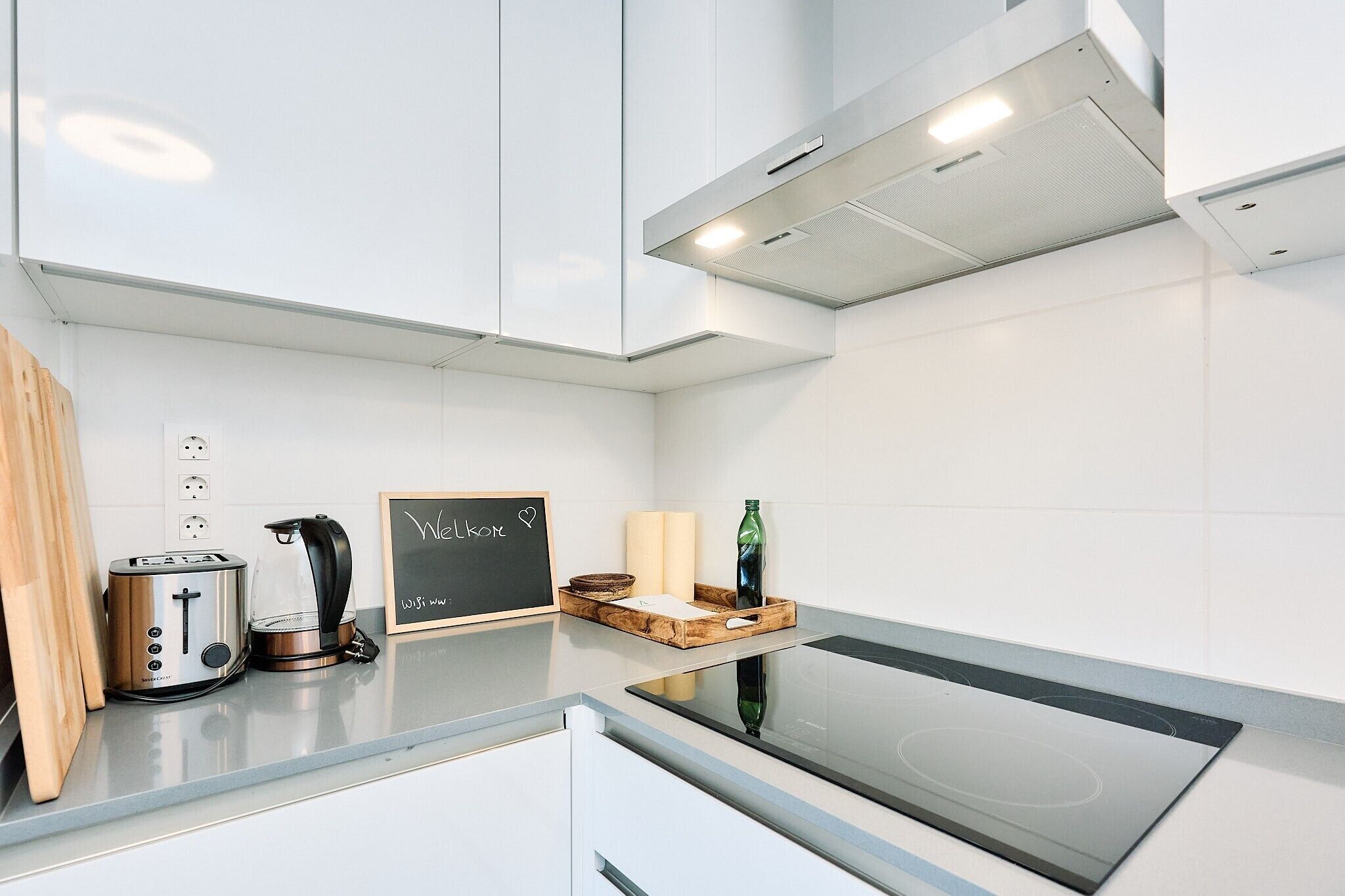 A modern kitchen with a toaster, kettle, and a chalkboard with 'Welcome' written on it.