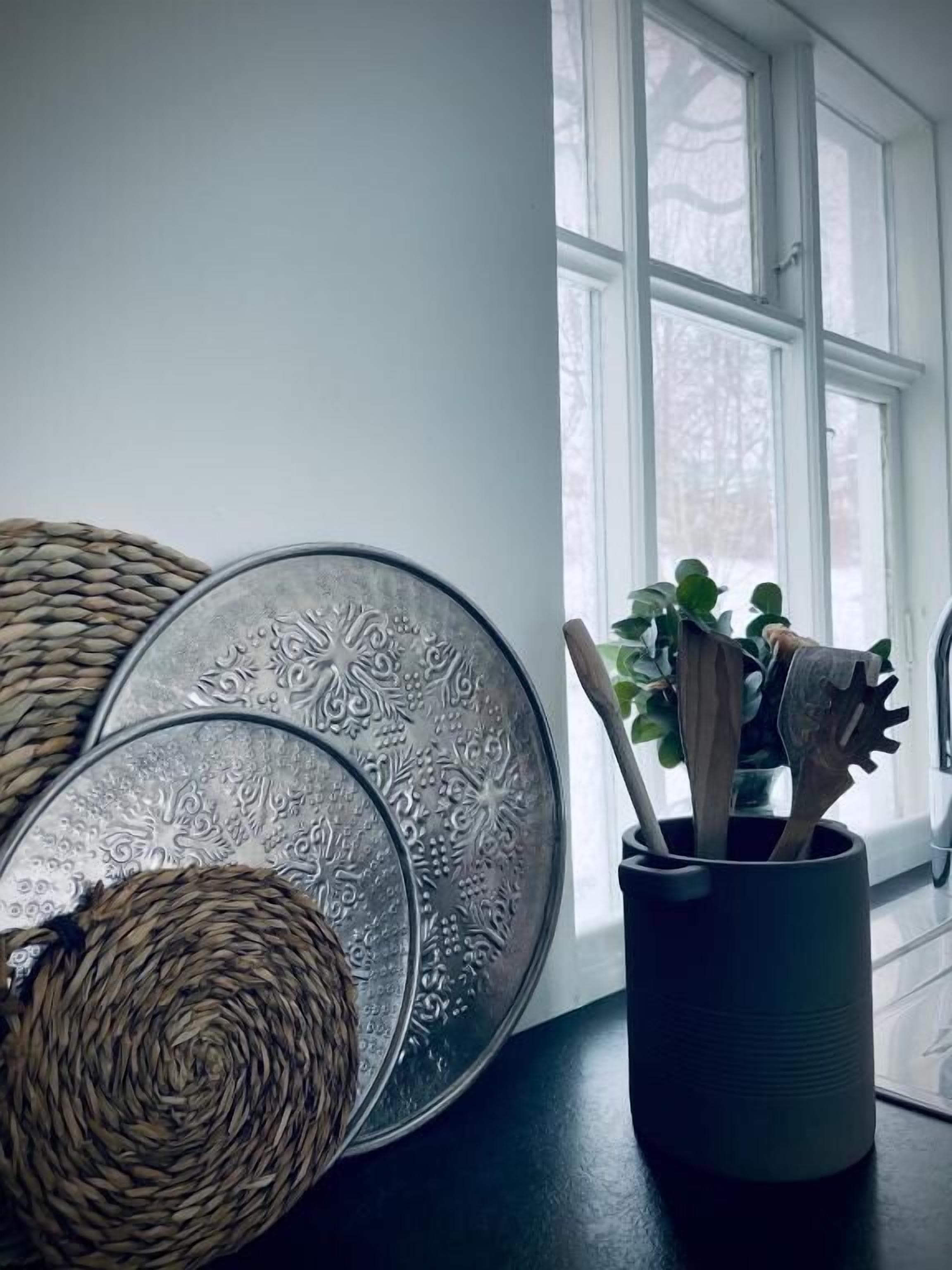 A room with a large window, a woven basket, a metal tray with intricate designs, and a wooden utensil holder with a plant.