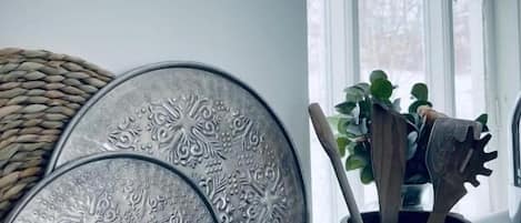 A room with a large window, a woven basket, a metal tray with intricate designs, and a wooden utensil holder with a plant.