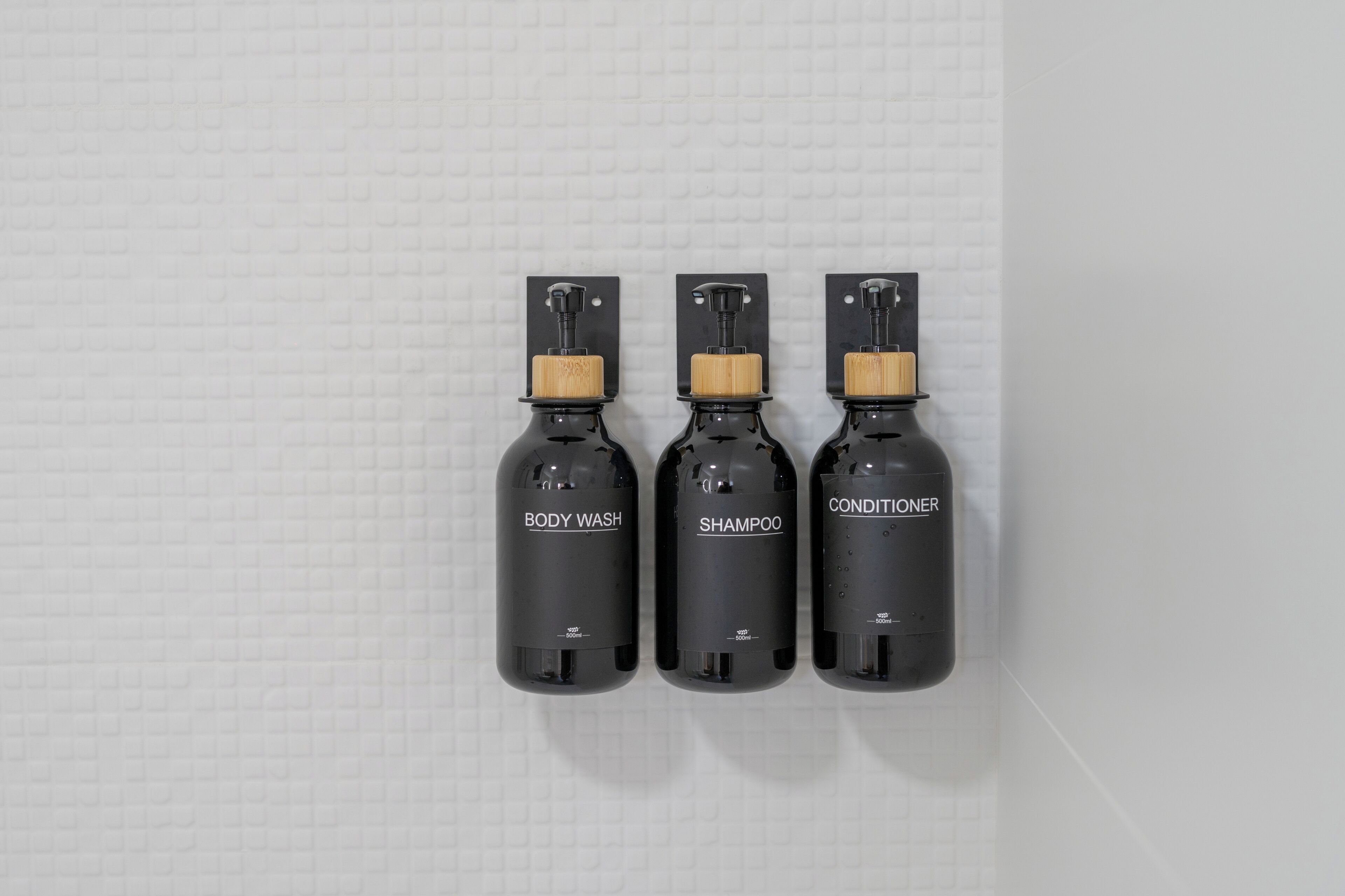 Tres botellas negras etiquetadas con JABÓN CORPORAL, CHAMPÚ y ACONDICIONADOR sujetas a una pared blanca con textura.