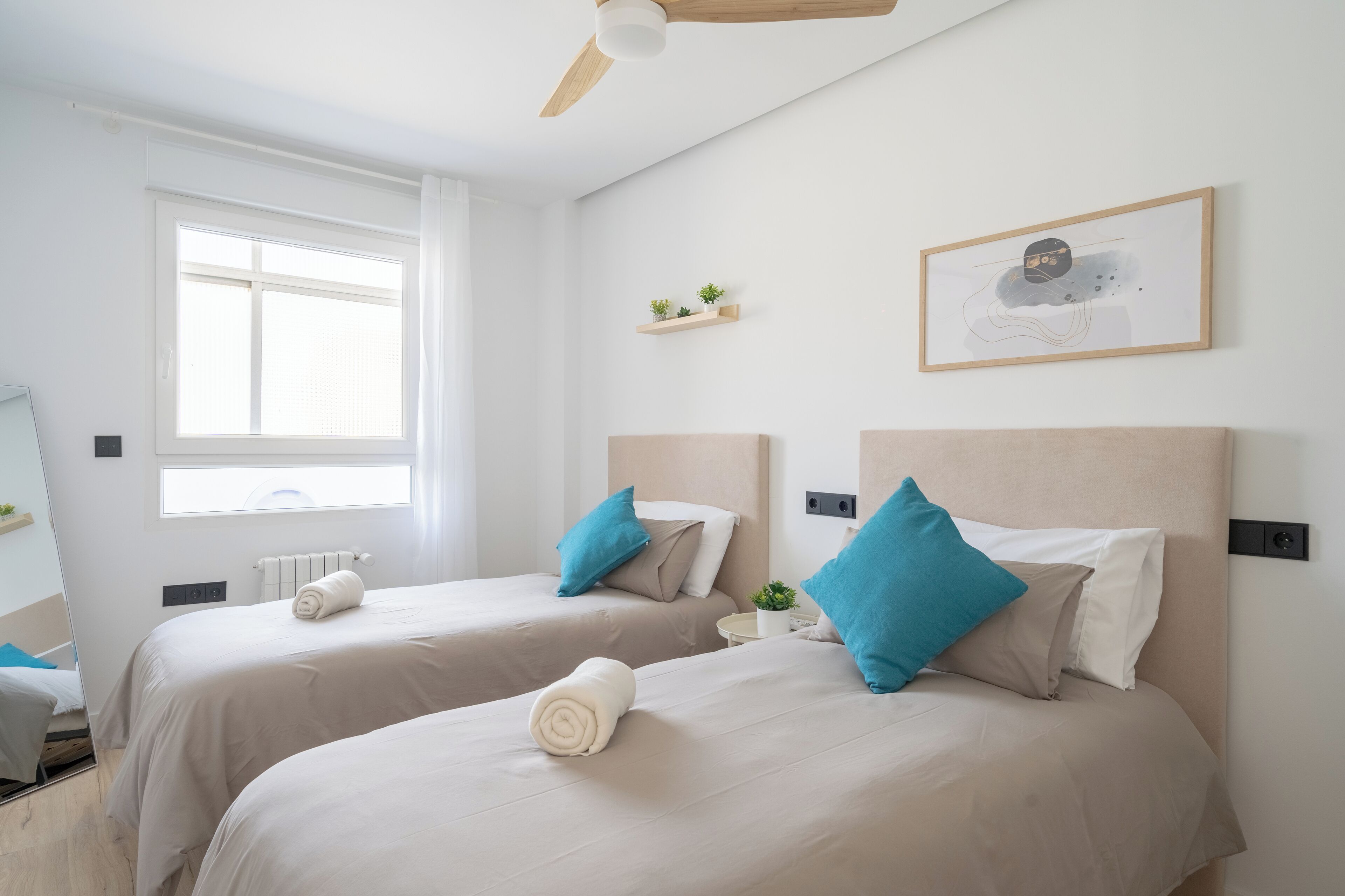 Une chambre d’hôtel avec deux lits, un ventilateur de plafond, une fenêtre, un radiateur, une petite plante et un tableau encadré au mur.