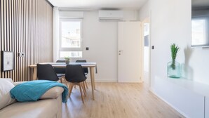 A modern living room with a sofa, a dining table with chairs, a wooden floor, and a white wall with a window.