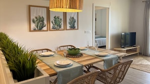 A modern dining area with a wooden table, chairs, and a central green bowl. There are framed cactus pictures on the wall, a television, and a door leading to another room. - Miramar Apartment (Denia)