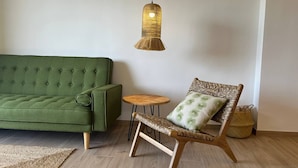 A room with a green sofa, a wicker chair with a cushion, a small round table, and a hanging light fixture. - Miramar Apartment (Denia)