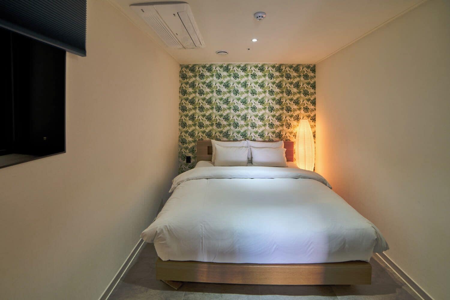 Une chambre d’hôtel étroite, avec un lit simple, un climatiseur fixé au mur et un papier peint à motifs.