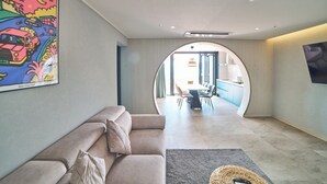 A modern living room with a sofa, a dining area with a table and chairs, and a kitchen visible through an archway.