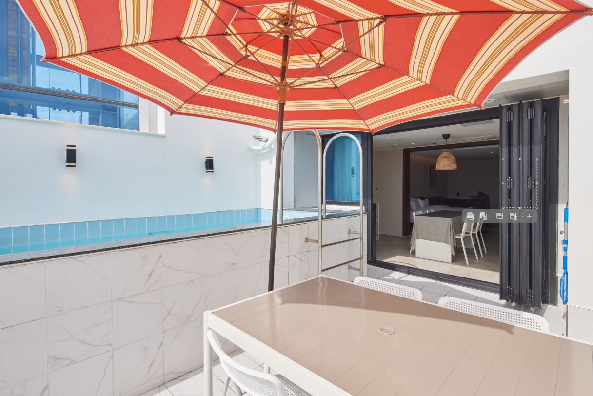 Zona junto a la piscina con mesa y sillas, un paraguas a rayas rojo y amarillo, y vistas a la piscina y habitaciones cercanas.