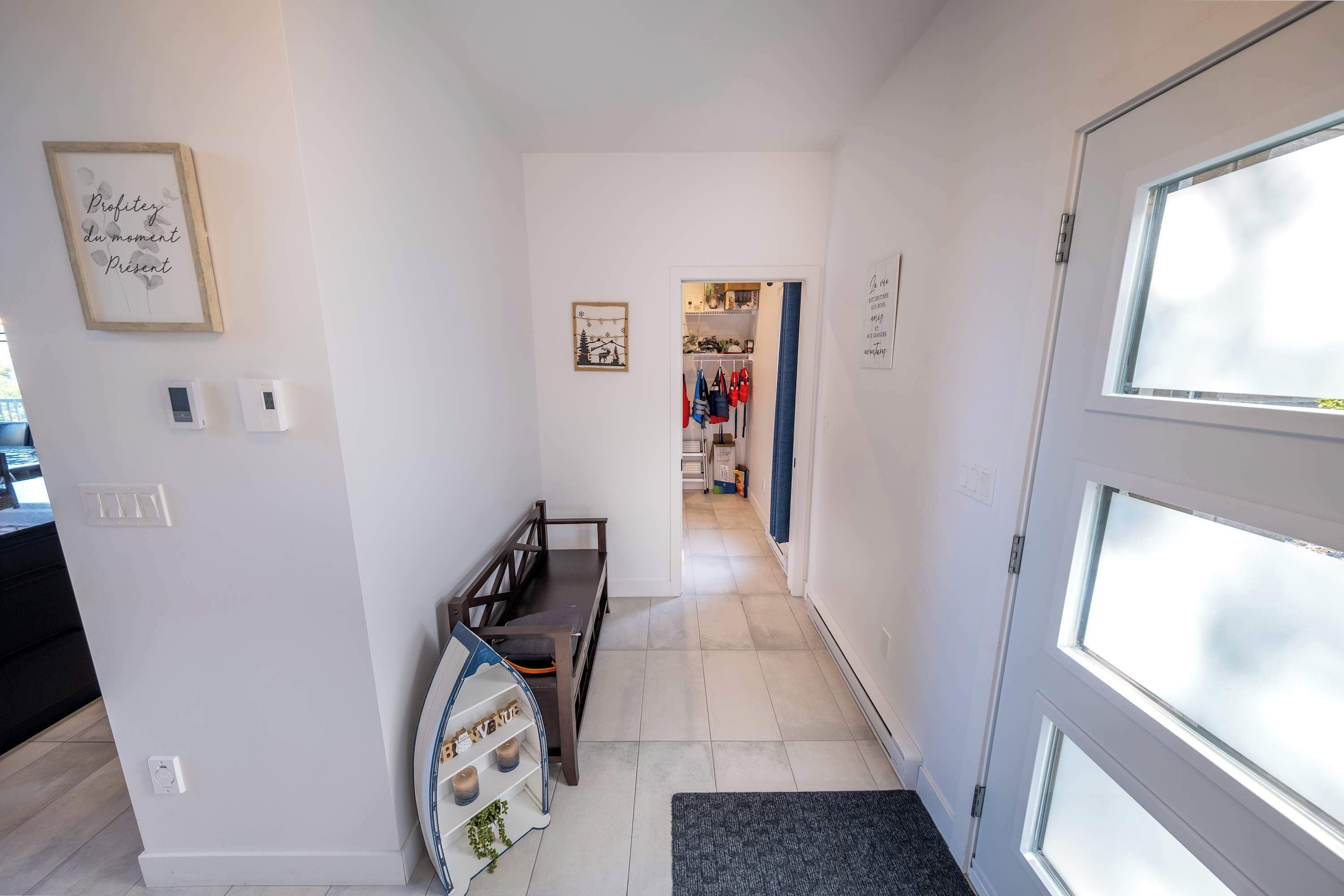 Een gang met een bankje, een klein plankje met decoratieve objecten, en een deur naar een kamer met een garderobe en een raam.
