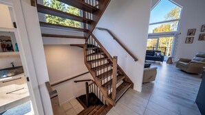 Ein modernes Interieur mit einer Holztreppe, einer Wohnbereits mit Sofa und Sesseln sowie einem großen Fenster mit Blick auf Bäume.