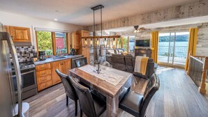 A modern kitchen with wooden cabinets, a dining area with a table and chairs, and a large window overlooking a lake. - SPA - Lake - Foosball Table | Chalet le Relaxarium (Lac-Etchemin)