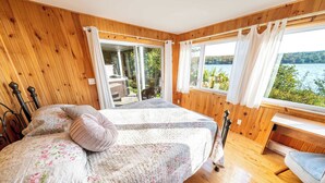 A bedroom with a wooden paneled wall, a bed with floral bedding, a desk, and a view of a lake through large windows. - SPA - Lake - Foosball Table | Chalet le Relaxarium (Lac-Etchemin)