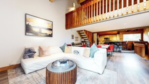 En moderne stue med en hvid hjørnesofa, et rundt sofabord og et spiseområde med røde stole. En trappe fører op til en etage med trægelændere.