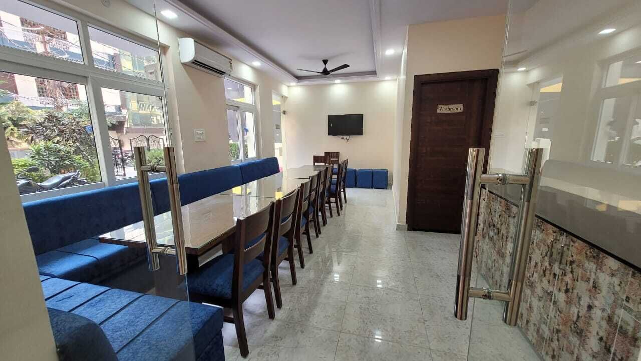 A hotel lobby with a long dining table, blue cushioned seating, a television on the wall, and a ceiling fan.