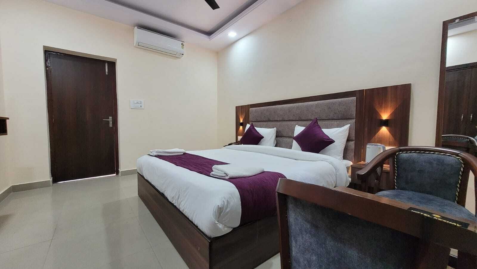 A hotel room with a double bed, a desk with a chair, a nightstand, and an air conditioning unit on the wall.
