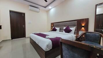 A hotel room with a double bed, a desk with a chair, a nightstand, and an air conditioning unit on the wall.