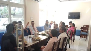 A group of people seated around a table in a room with a large window, a flat-screen TV mounted on the wall, and a sofa in the background.