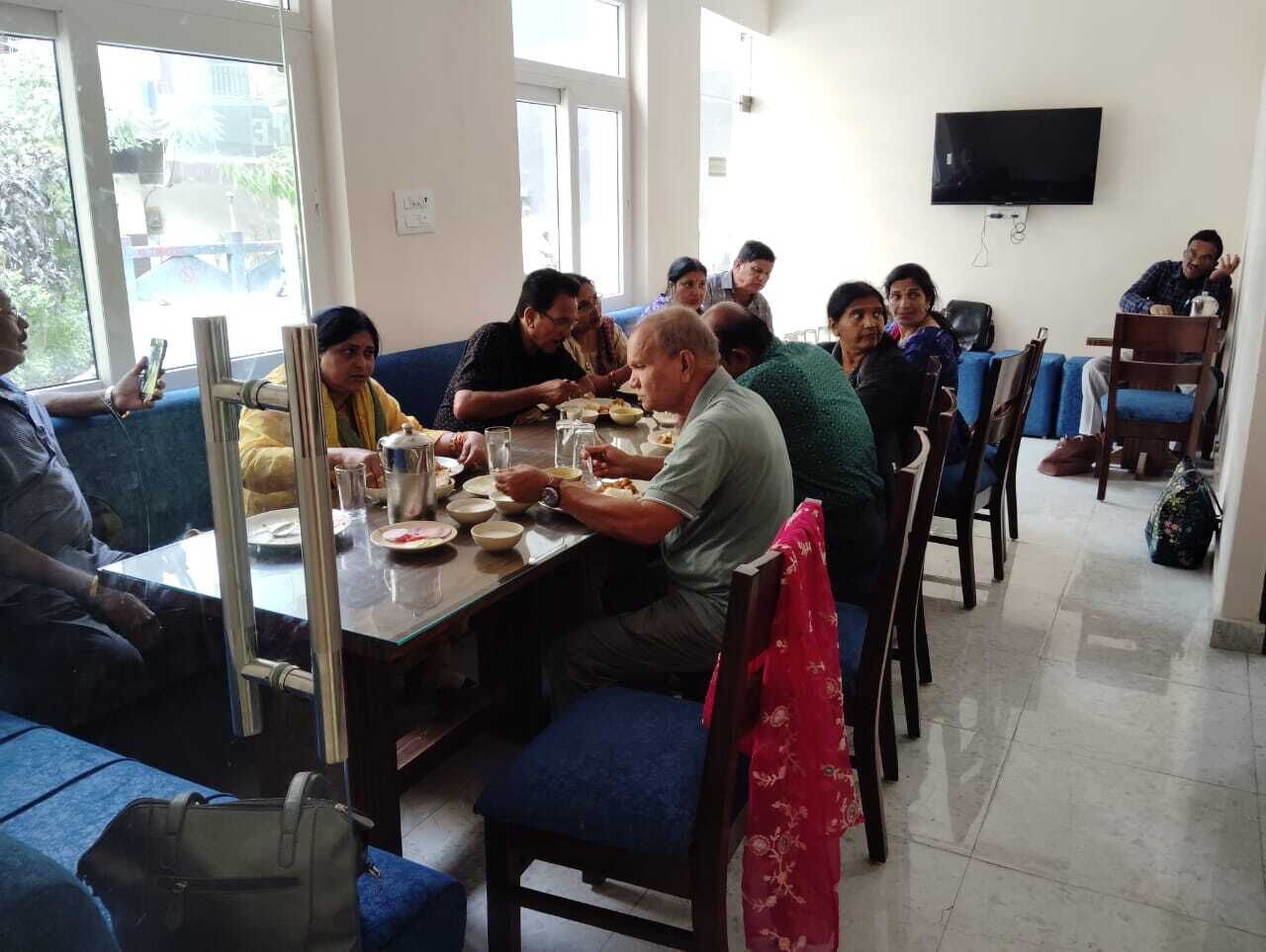 A group of people dining in a room with a large window, a flat-screen TV mounted on the wall, and a blue sofa in the foreground.