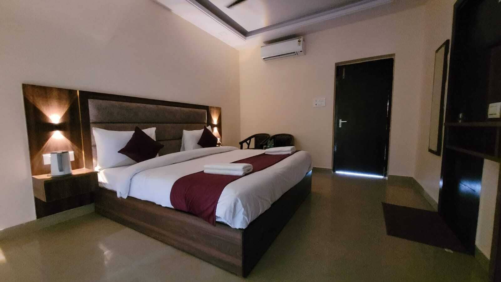 Una camera d'albergo con un letto grande, un comodino con una lampada, un condizionatore d'aria a parete e una porta che dà su un'altra stanza.