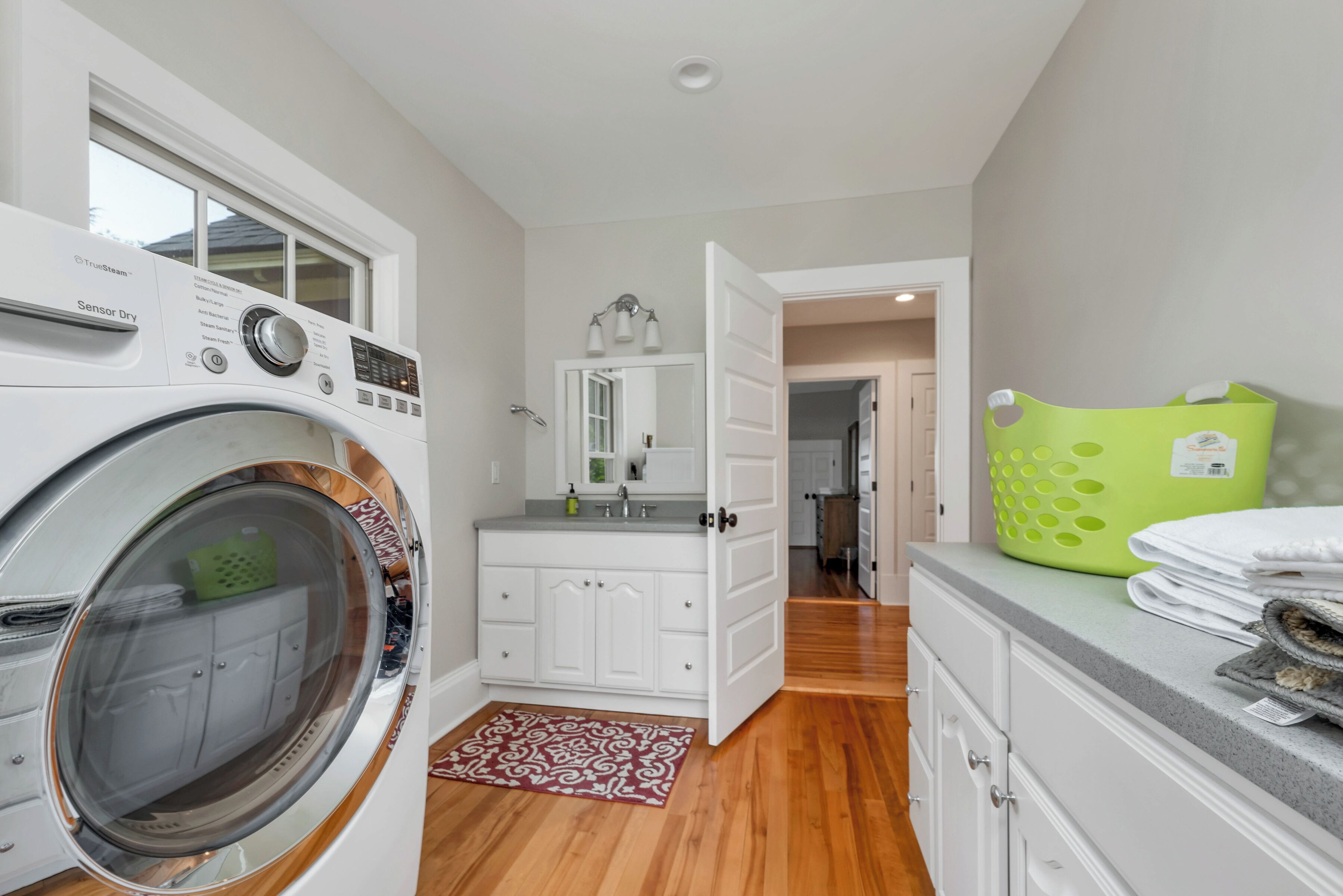 Une buanderie équipée d’un lave-linge à chargement frontal, d’un évier, d’un miroir et d’un panier à linge vert.