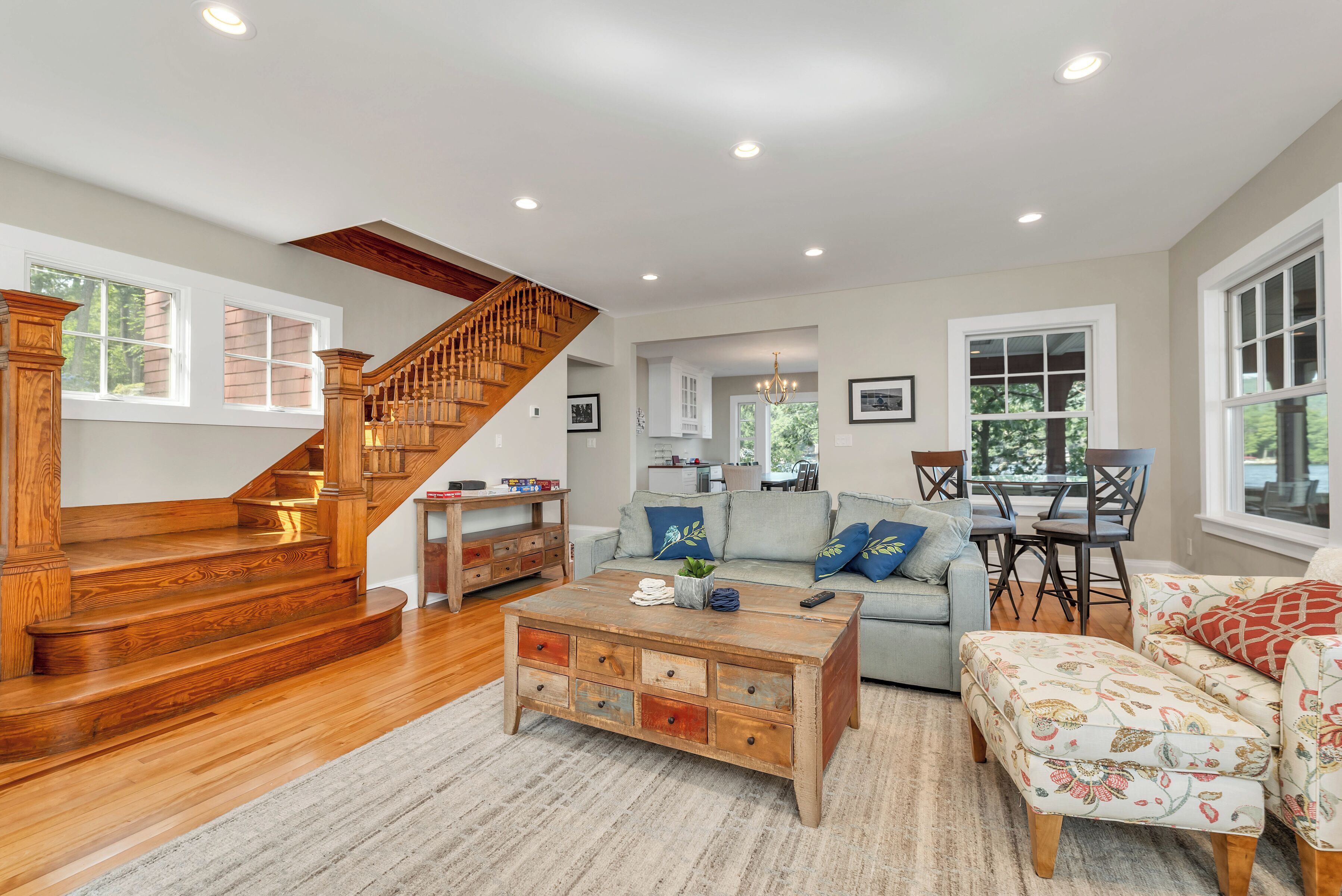 A spacious living room with a wooden staircase, a sofa set, a coffee table, and a dining area with a table and chairs.