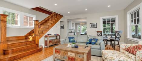 A spacious living room with a wooden staircase, a sofa set, a coffee table, and a dining area with a table and chairs.