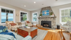 A living room with a stone fireplace, large windows, and a view of the outdoors. - Lake Winnipesauke sleeps 9 with stunning sunset views (Alton Bay)
