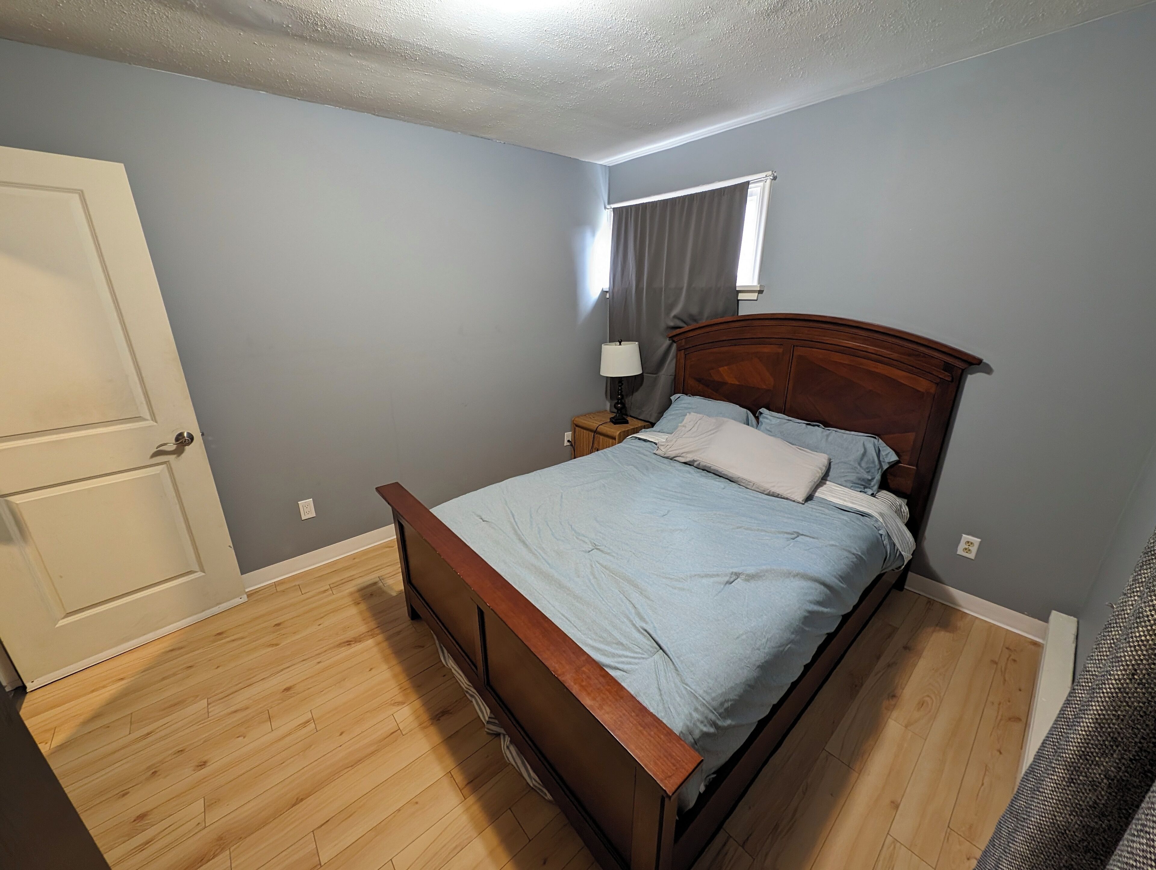 Een slaapkamer met een houten bed, een nachtkastje, een raam met gordijnen en een deur.