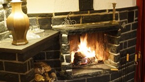 A stone fireplace with a fire burning, a metal shelf with a lamp, and a basket of firewood. - Exclusive Group House in Winterberg in the Forest (Winterberg)