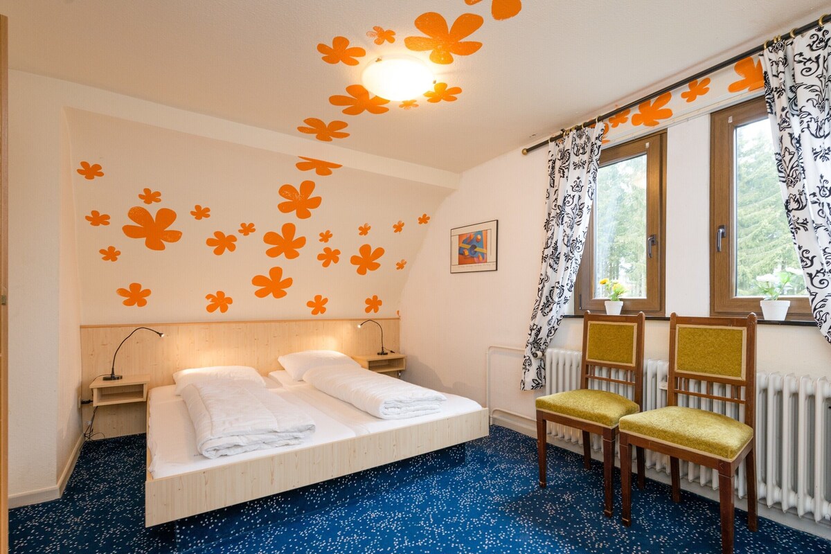 A bedroom with a bed, two bedside tables, a nightstand, two chairs, a radiator, a window with curtains, and a ceiling light.