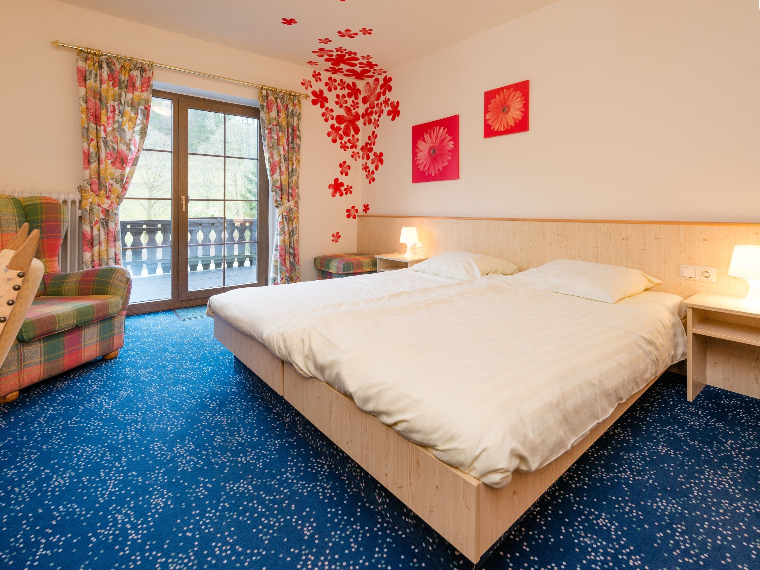 Ein Hotelzimmer mit einem großen Bett, zwei Nachttischen, einem Sessel und Blick ins Freie durch eine Schiebetür.