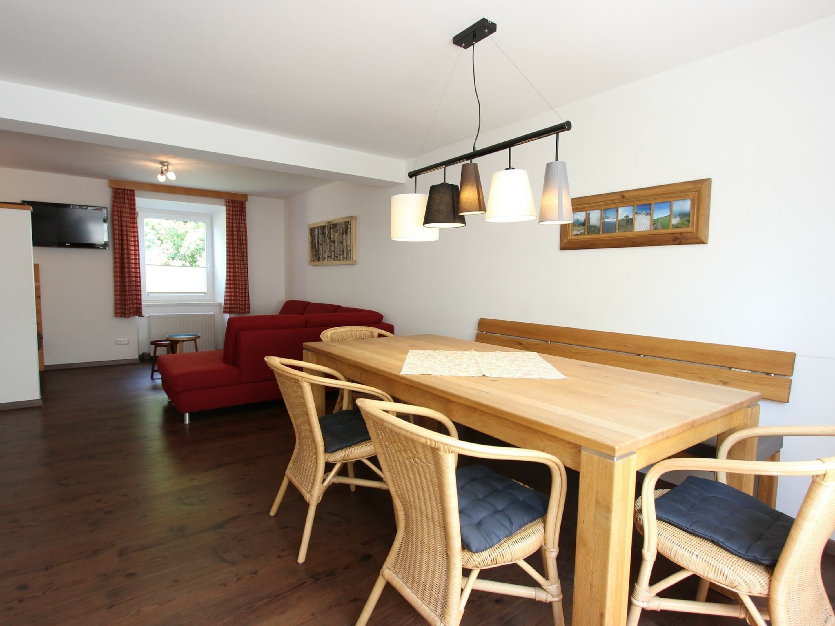 A dining area with a wooden table and wicker chairs, a red sofa, and a television mounted on the wall.