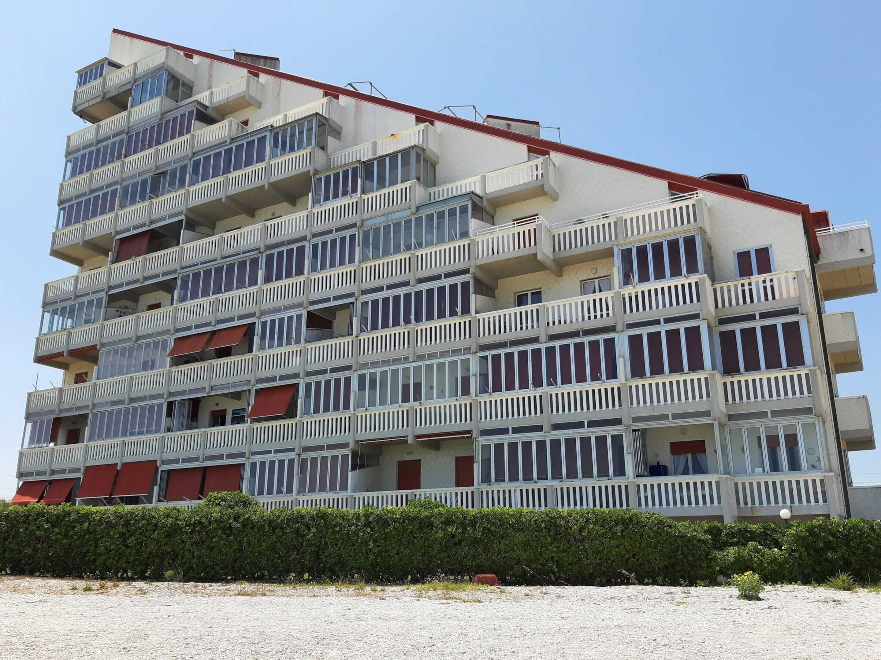 A multi-story building with balconies, a red roof, and a gravel ground.