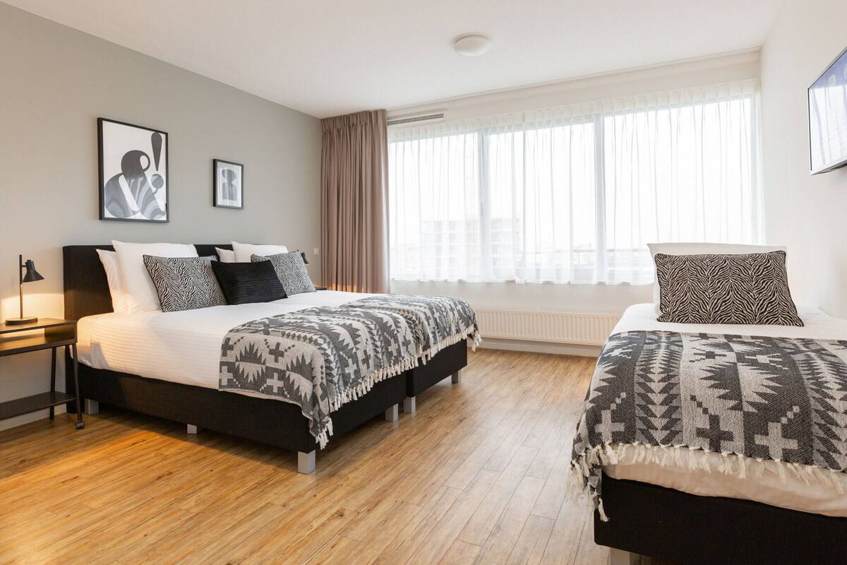 A modern hotel room with two beds, a large window with sheer curtains, and a minimalist design.