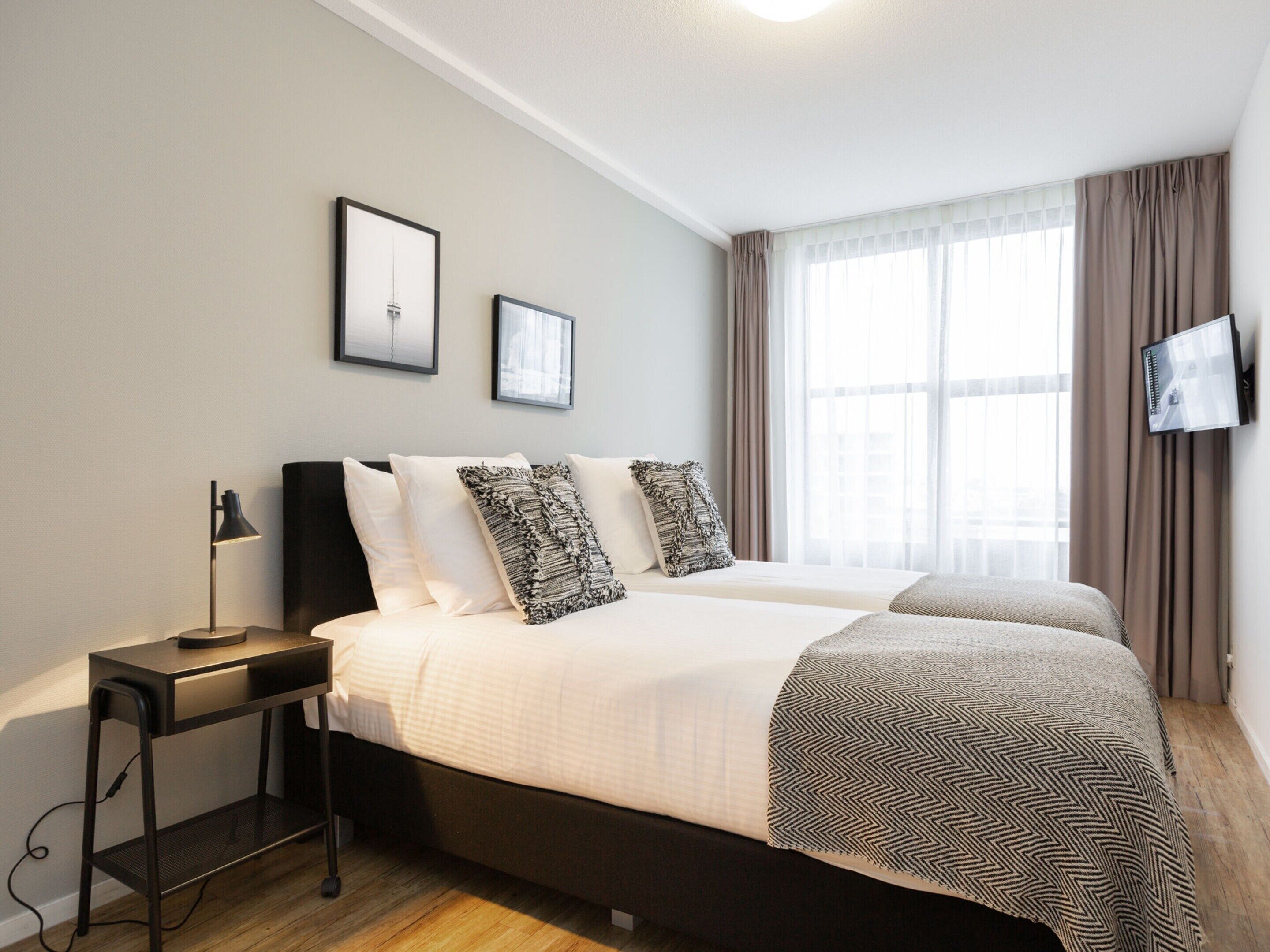 A double bed with a black frame, two pillows, and a patterned blanket. There is a bedside table with a lamp and a TV mounted on the wall.