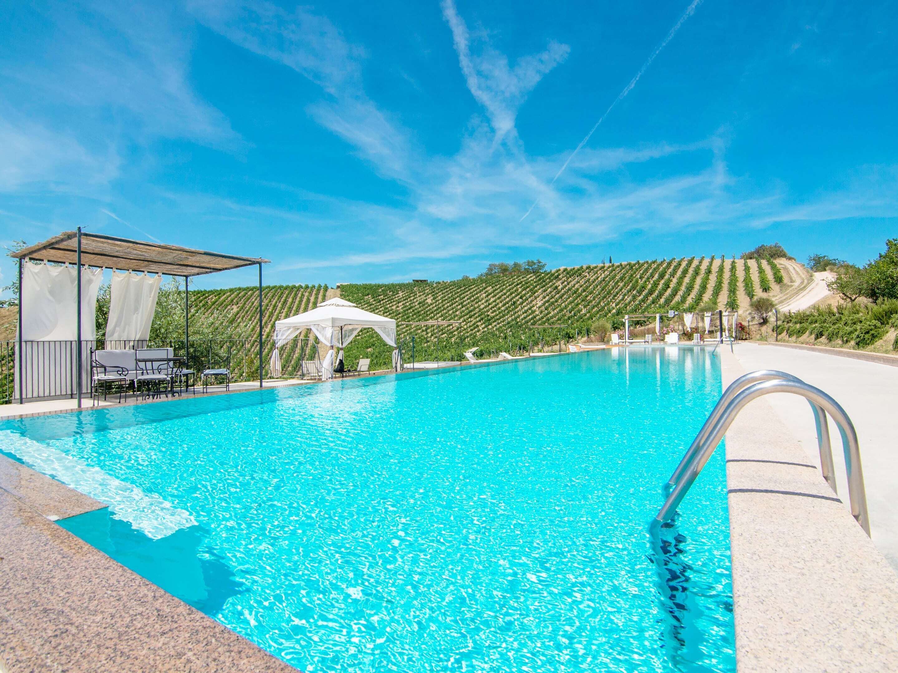 Ein Swimmingpool mit klarem, blauem Wasser, umgeben von einer Pergola und Liegestühlen, mit einem Weinberg im Hintergrund.