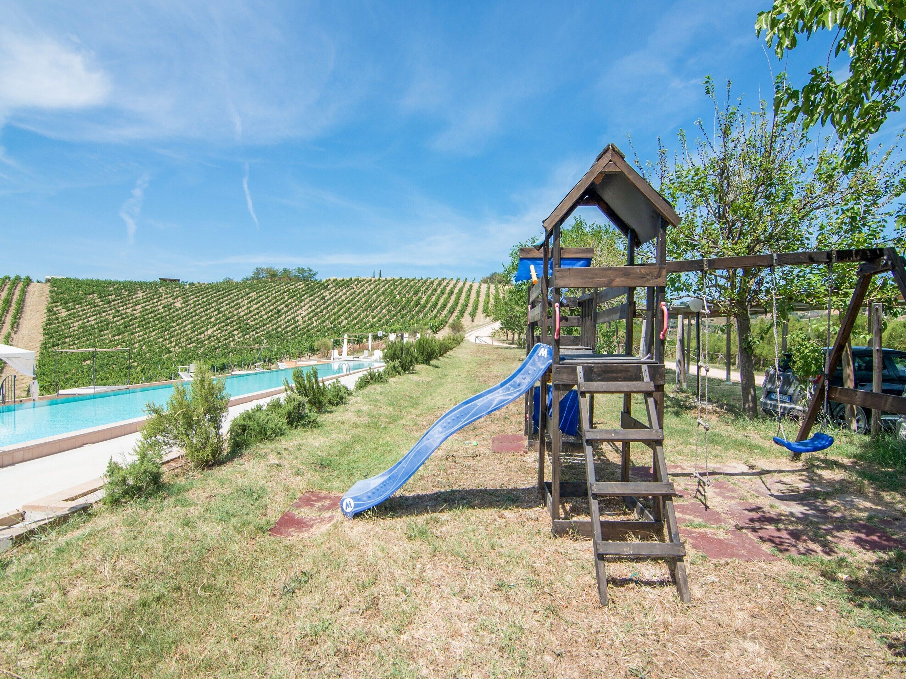 Um parquinho com escorrega e estrutura de escalada, piscina e fileiras de vinhedos ao fundo.