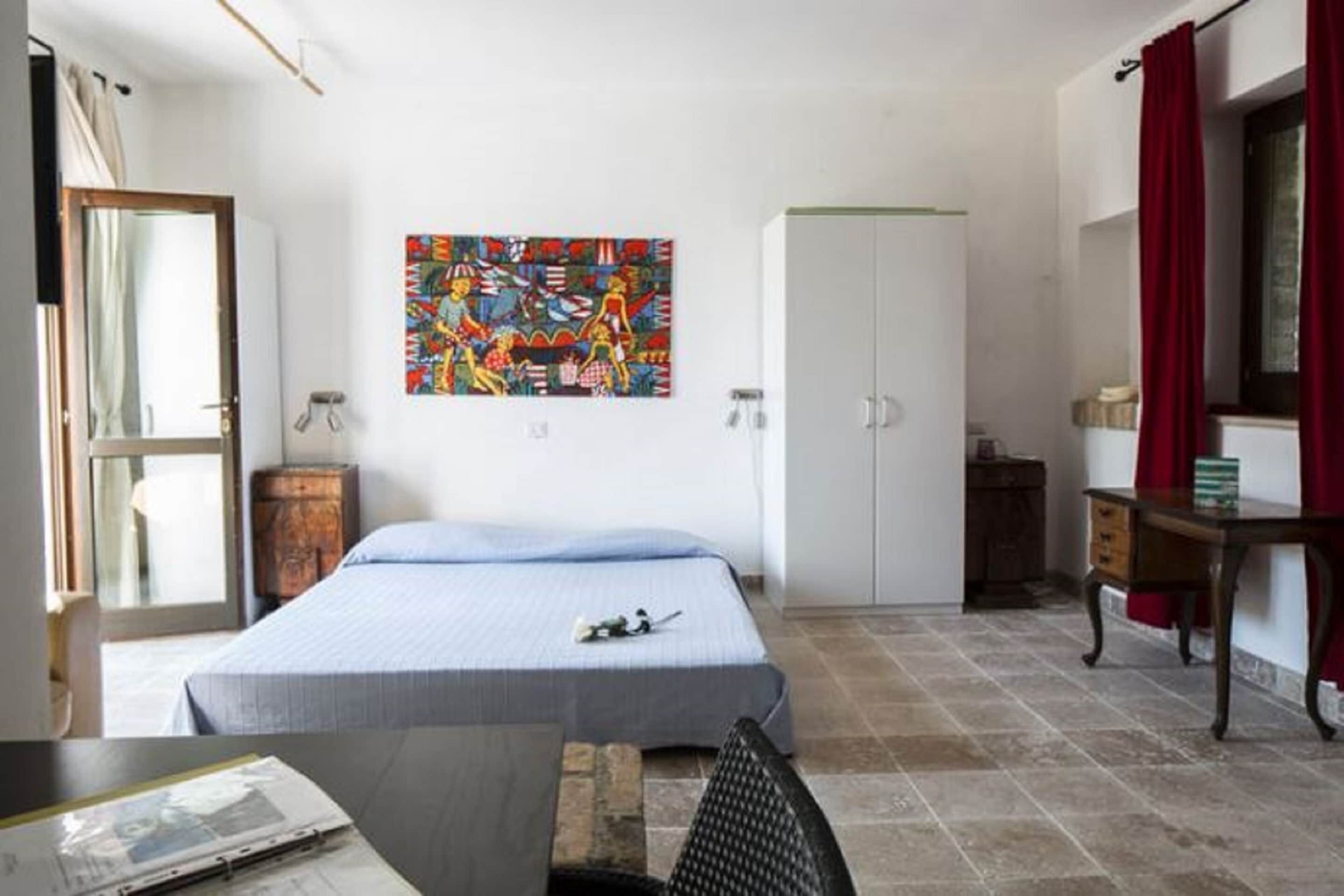 A bedroom with a bed, a desk, a chair, a wardrobe, and a colorful abstract painting on the wall.