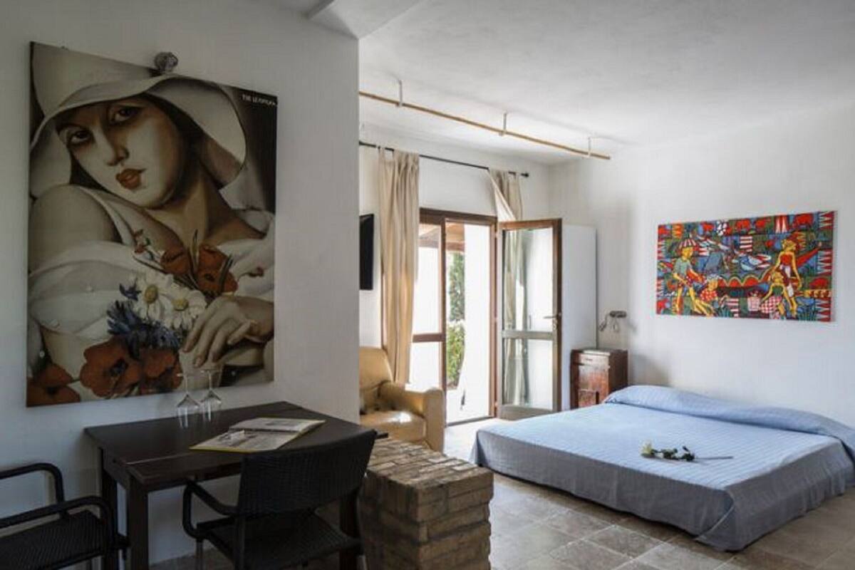 Une pièce comprenant un lit, une table à manger avec des chaises, un grand tableau au mur et une fenêtre avec des rideaux.