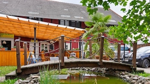 A wooden bridge over a pond with rocks and plants, a restaurant with outdoor seating, and a building with a tiled roof.