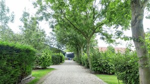 Ein Kiesweg, der von dichten, grünen Hecken und Bäumen gesäumt wird und zu einem Gebäude mit rotem Dach führt.