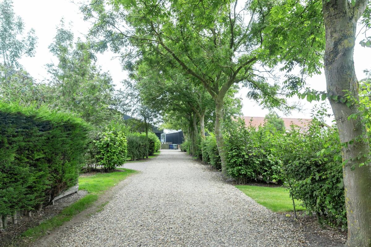 Een grindpad, begrensd door dichte groene hagen en bomen, leidt naar een gebouw met een rood dak.