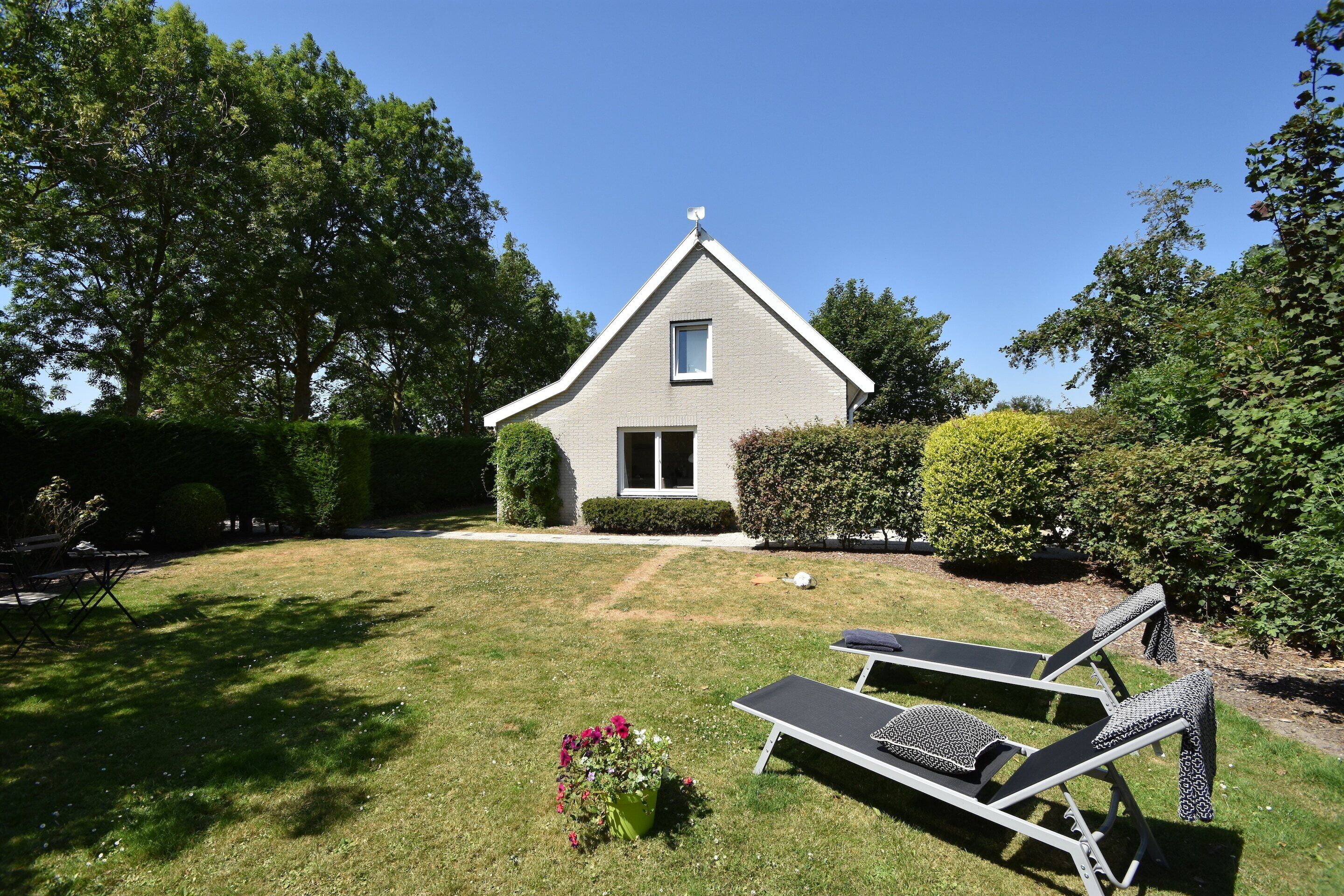 A house with a triangular roof, a small window, and a door. There is a lawn with two lounge chairs and a potted plant.