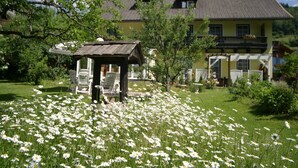 A house with a garden full of white flowers, a wooden gazebo, and a large yellow building in the background. - Cozy Apartment in Feld am See Near Ski Slopes (Feld am See)