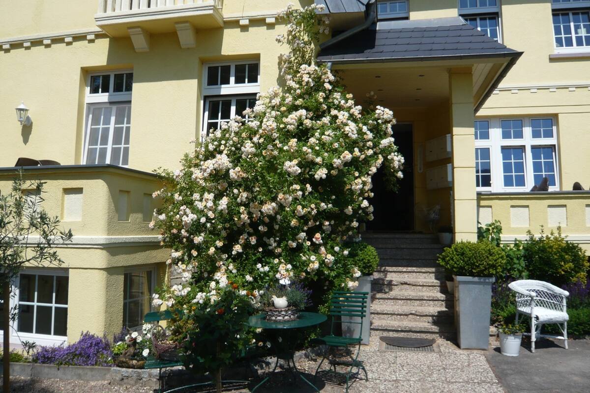 A yellow building with a balcony, a rose bush, a table with chairs, and a chair.